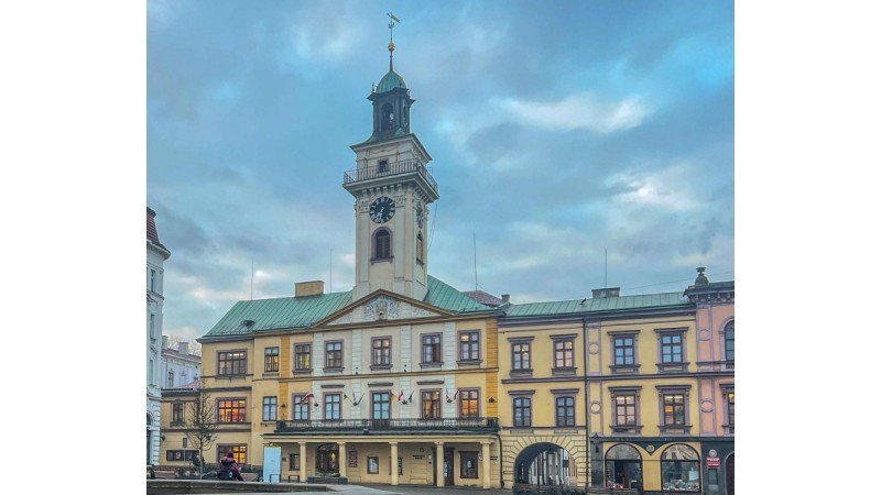 Rynek z ratuszem. Jasny budynek z wieżą i zegarem, na dole podcienia. Przed nim pusty plac.